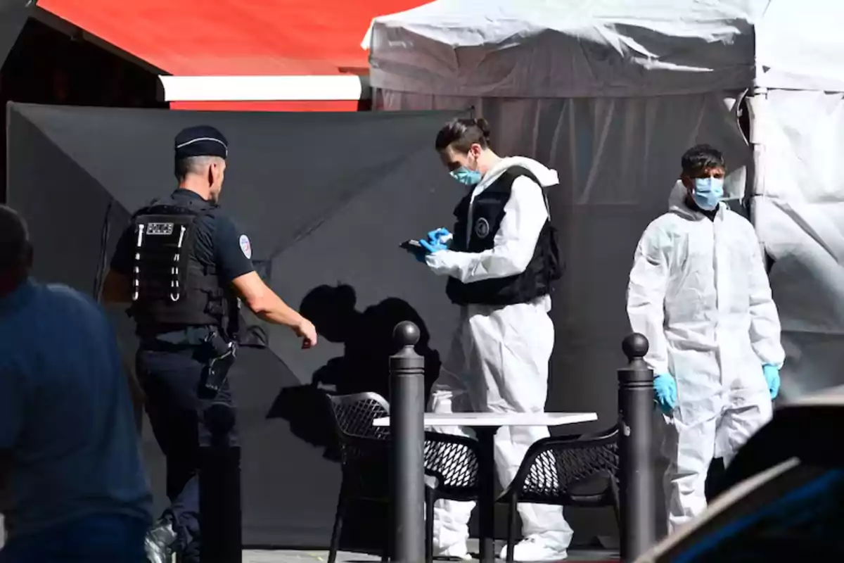 Police and forensic personnel work at a cordoned-off outdoor scene next to a table and chairs, wearing gloves and masks.
