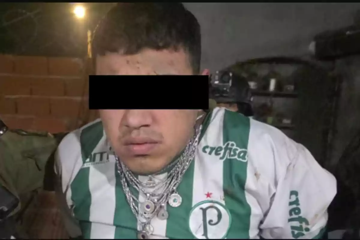 Man with his eyes covered by a black bar, wearing a green and white soccer jersey and several silver chains, in an indoor setting with brick walls.