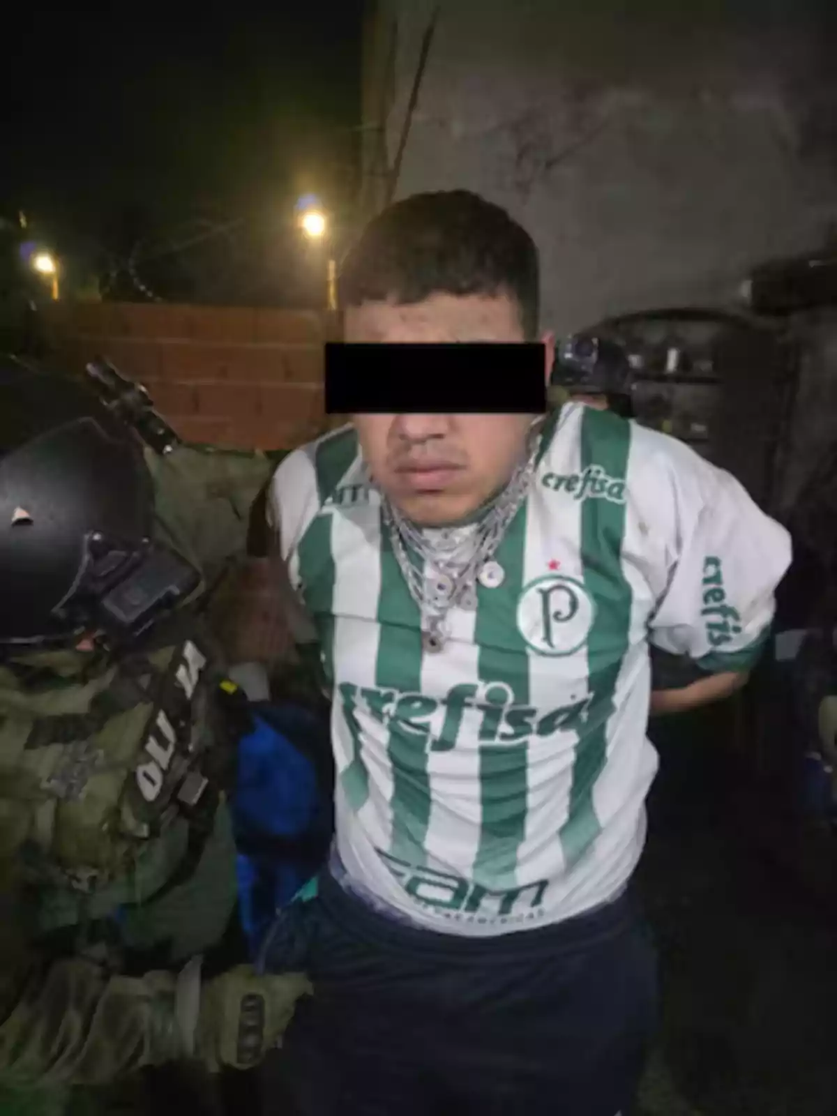 Man in a green and white soccer jersey being detained by a police officer at night with his eyes covered by a black bar Man in a green and white soccer jersey being detained by a police officer at night with his eyes covered by a black bar