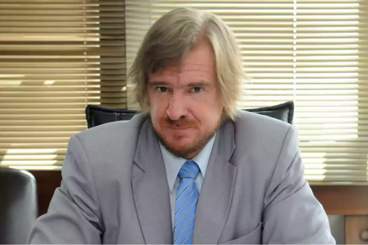 Blond-haired, bearded man sitting in an office wearing a gray suit and blue tie with blinds in the background Blond-haired, bearded man sitting in an office wearing a gray suit and blue tie with blinds in the background