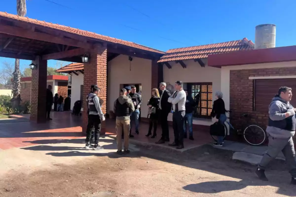 Several people, including police officers and civilians, are gathered in front of a brick house with a red tile roof on a sunny day. Several people, including police officers and civilians, are gathered in front of a brick house with a red tile roof on a sunny day.