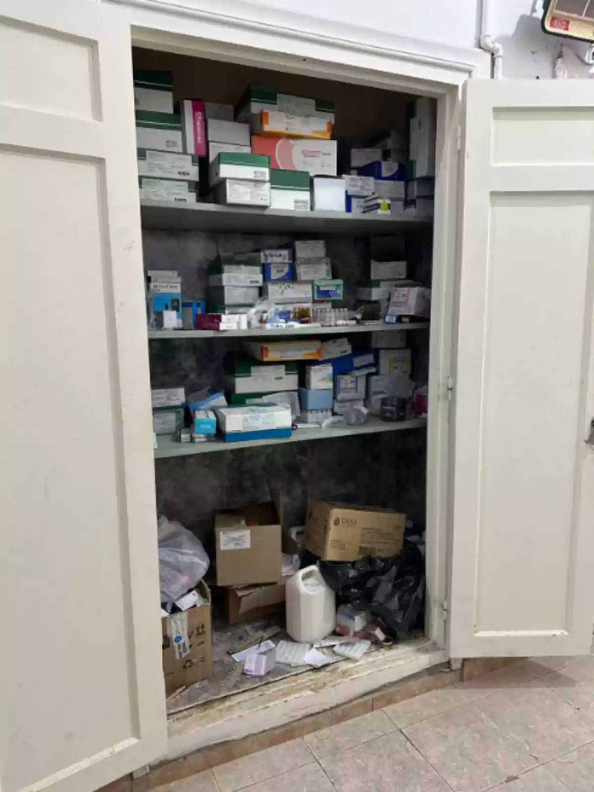 Open white cabinet with shelves full of boxes and disorganized medications, as well as boxes and containers on the floor Open white cabinet with shelves full of boxes and disorganized medications, as well as boxes and containers on the floor