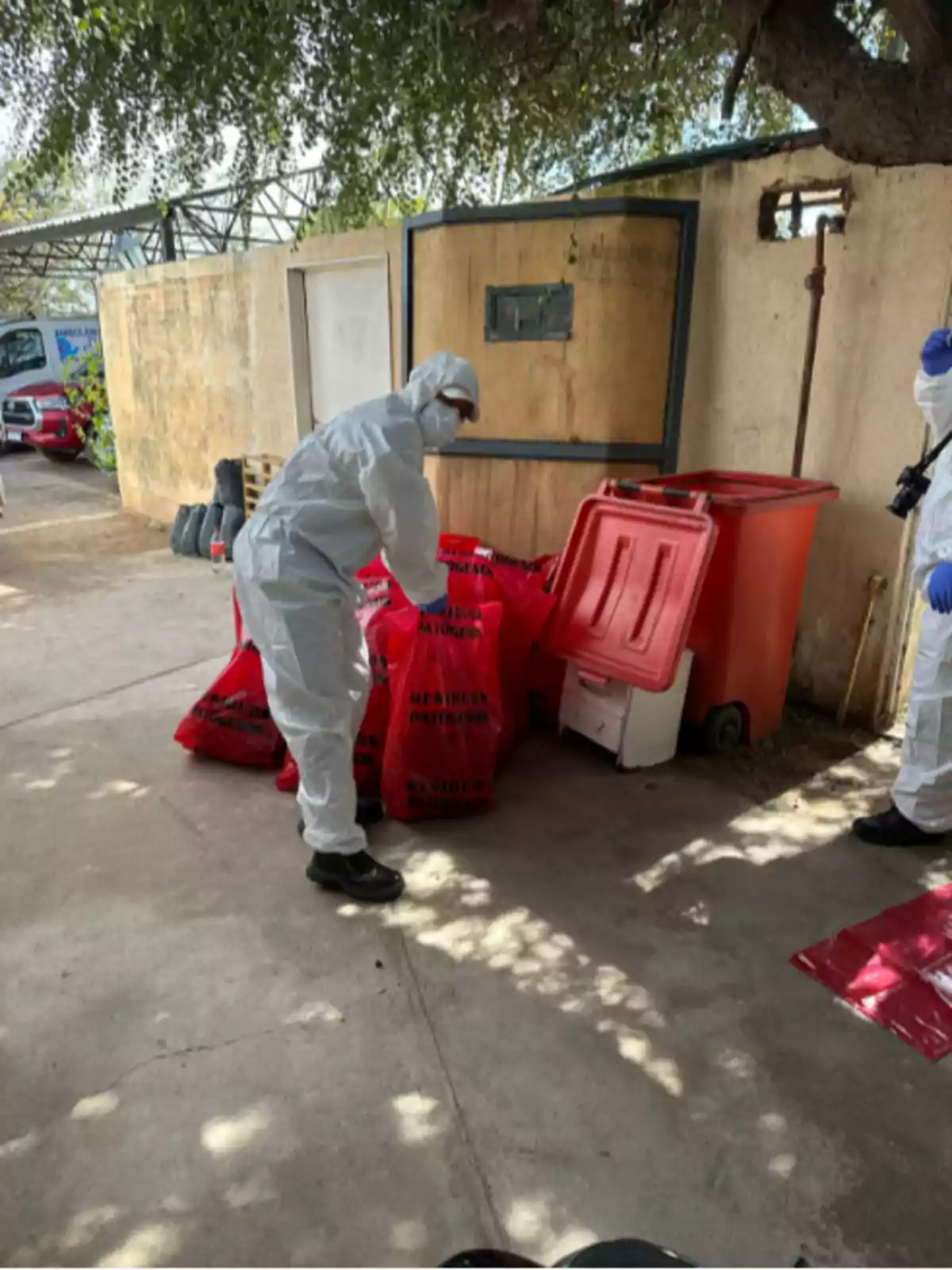 Person wearing protective suit handling red hazardous waste bags next to a container in an outdoor area Person wearing protective suit handling red hazardous waste bags next to a container in an outdoor area