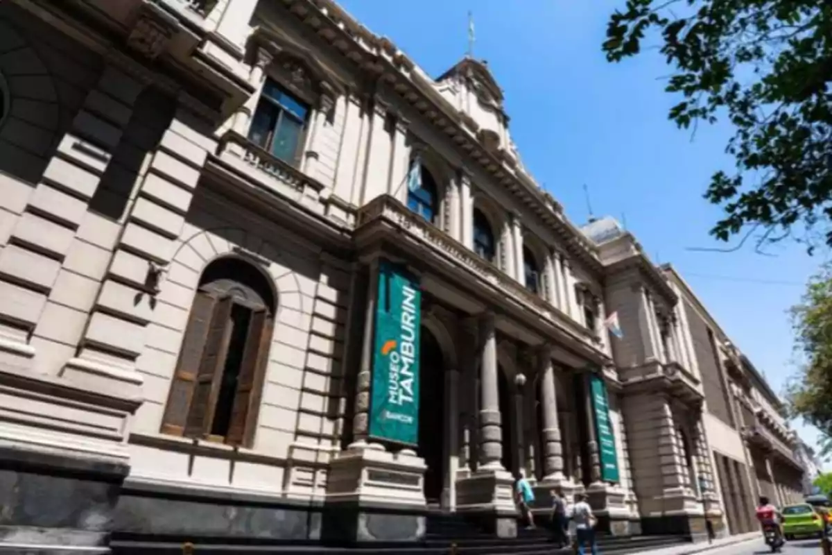 Fachada de un edificio histórico con columnas y ventanas grandes, donde se observan personas subiendo las escaleras de entrada y un letrero que indica Museo Tamburini bajo un cielo despejado