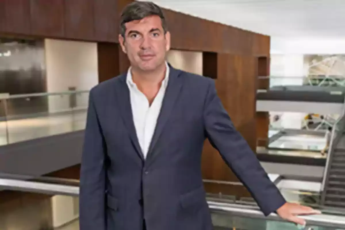 Hombre de cabello corto y oscuro con traje azul y camisa blanca posando en el interior de un edificio moderno con barandales de cristal y paredes de madera Hombre de cabello corto y oscuro con traje azul y camisa blanca posando en el interior de un edificio moderno con barandales de cristal y paredes de madera