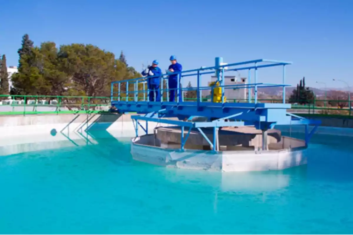 Two workers wearing helmets and blue uniforms inspect a water treatment plant under a clear sky.