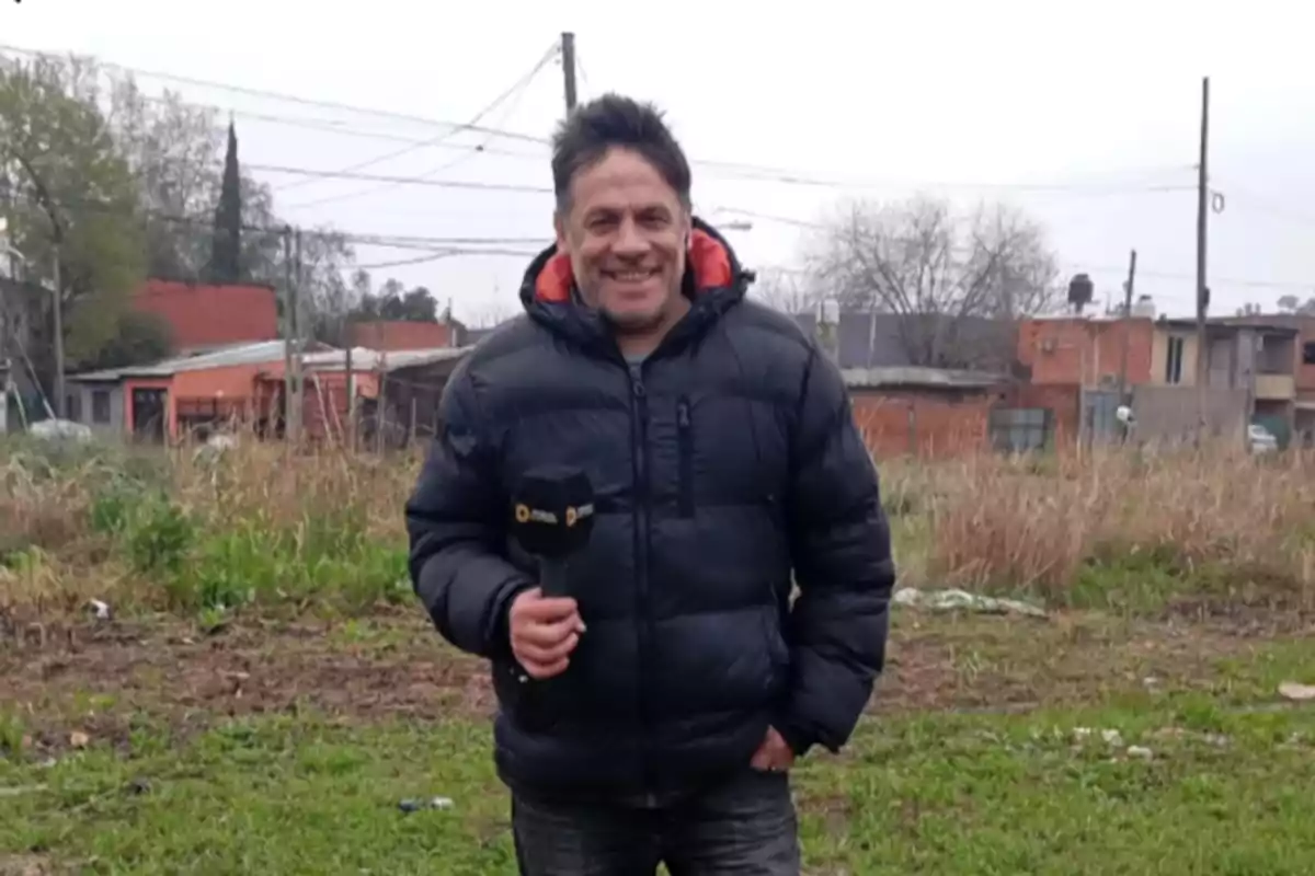 Man wearing a black jacket holding a microphone on a grassy field with houses in the background Man wearing a black jacket holding a microphone on a grassy field with houses in the background