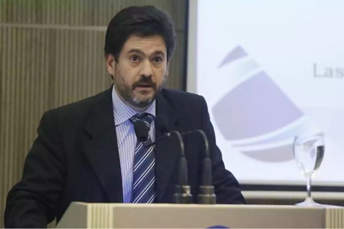 Man in a suit speaking into a microphone at a podium with a presentation screen in the background