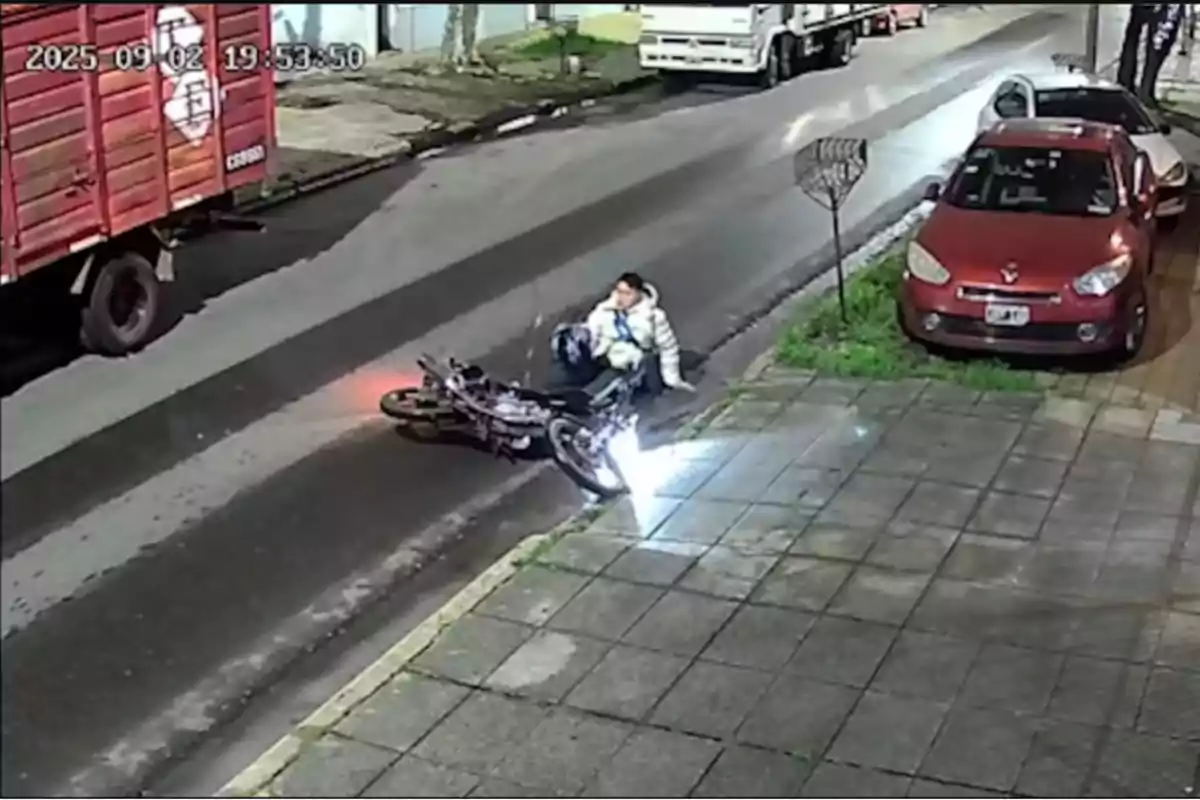 A man in a white jacket is sitting on the ground next to a fallen motorcycle on a wet street at night, near a sidewalk and several parked cars. A man in a white jacket is sitting on the ground next to a fallen motorcycle on a wet street at night, near a sidewalk and several parked cars.