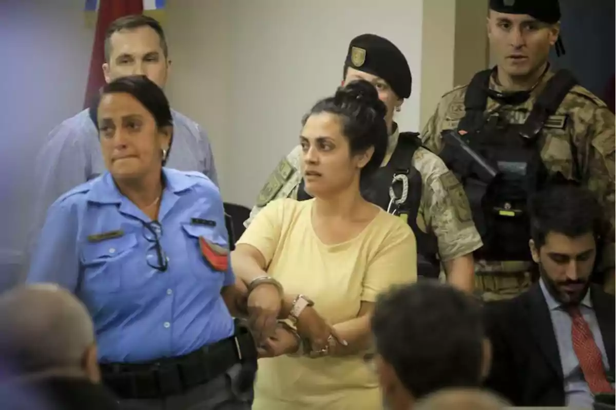 A handcuffed woman is escorted by a police officer while several officers and people watch in a room.