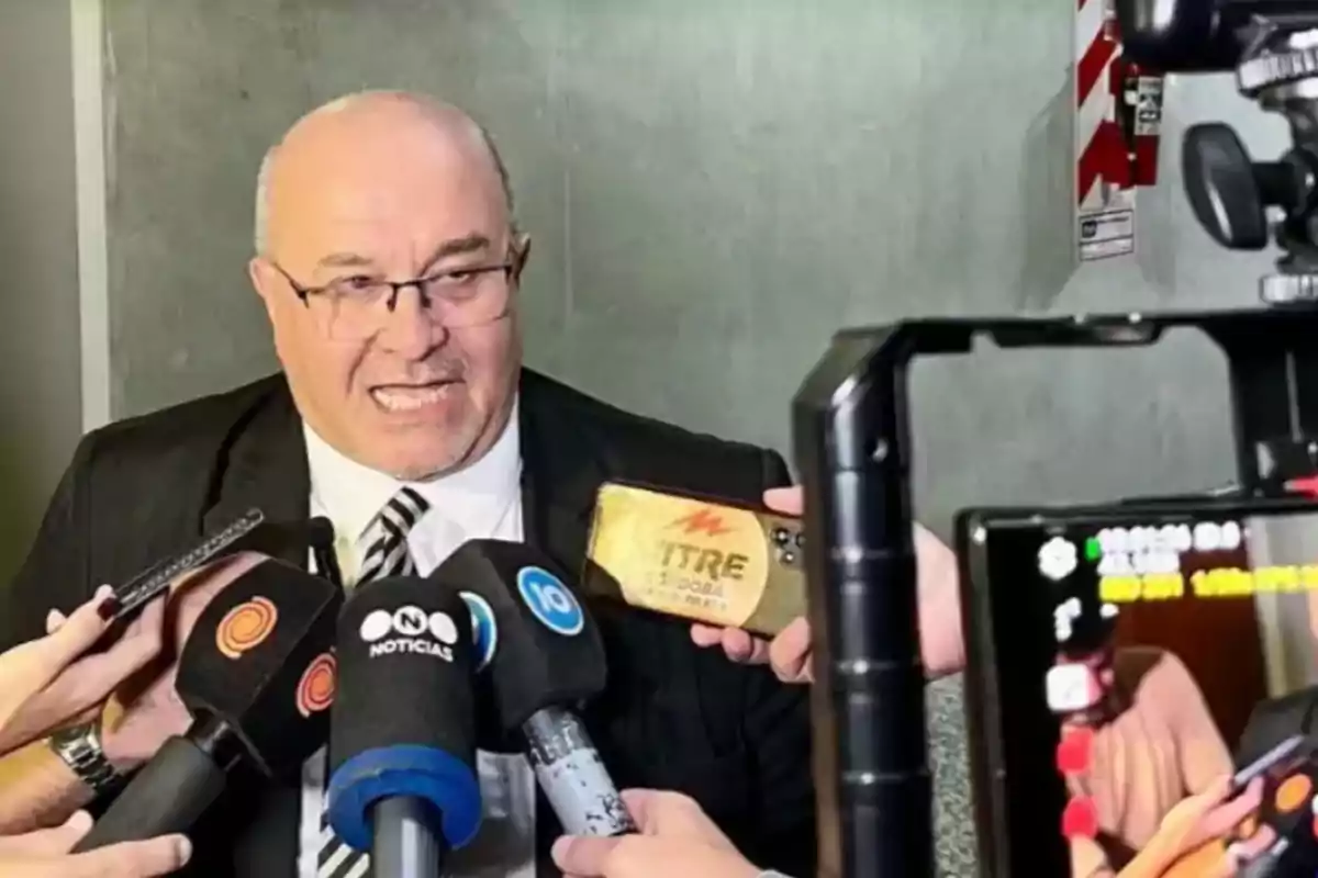 Man in a suit and tie giving statements to several media outlets in front of microphones and cameras Man in a suit and tie giving statements to several media outlets in front of microphones and cameras