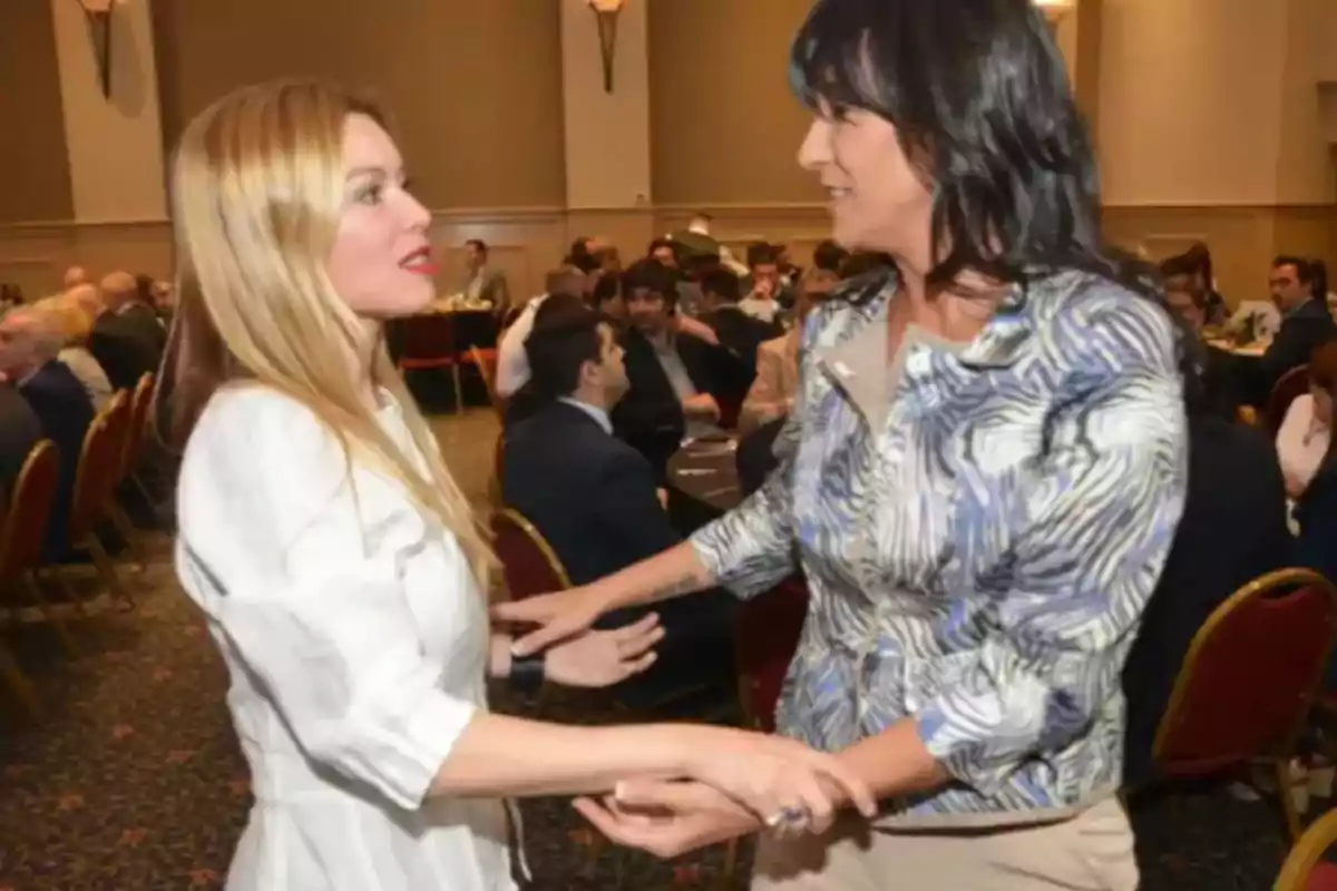 Two women greet each other while standing in a room full of people seated at round tables.