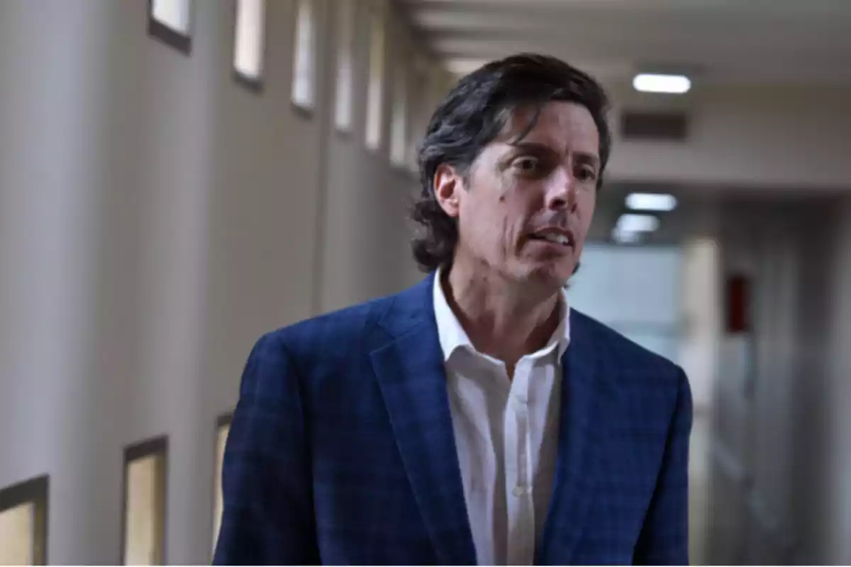 Hombre de cabello oscuro y ondulado vistiendo saco azul y camisa blanca en un pasillo iluminado