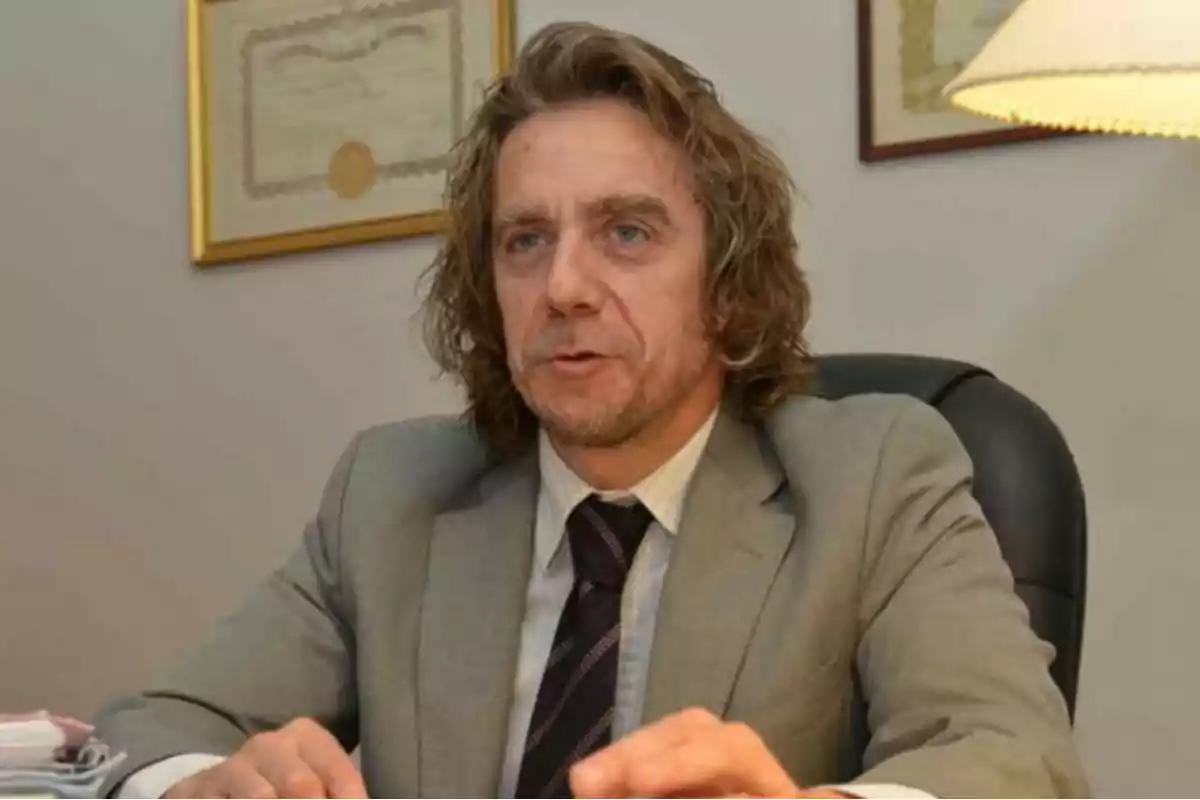 Hombre de cabello largo y rubio con traje gris sentado en una oficina frente a un escritorio Hombre de cabello largo y rubio con traje gris sentado en una oficina frente a un escritorio