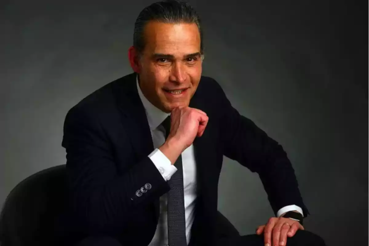 Middle-aged man in a dark suit and gray tie posing with a smile, hand resting on his chin, against a gray background Middle-aged man in a dark suit and gray tie posing with a smile, hand resting on his chin, against a gray background