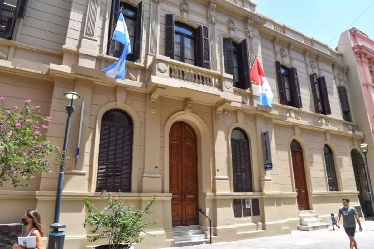 Facade of an old two-story building with wooden windows, large doors, two flags, and people walking on the sidewalk on a sunny day.