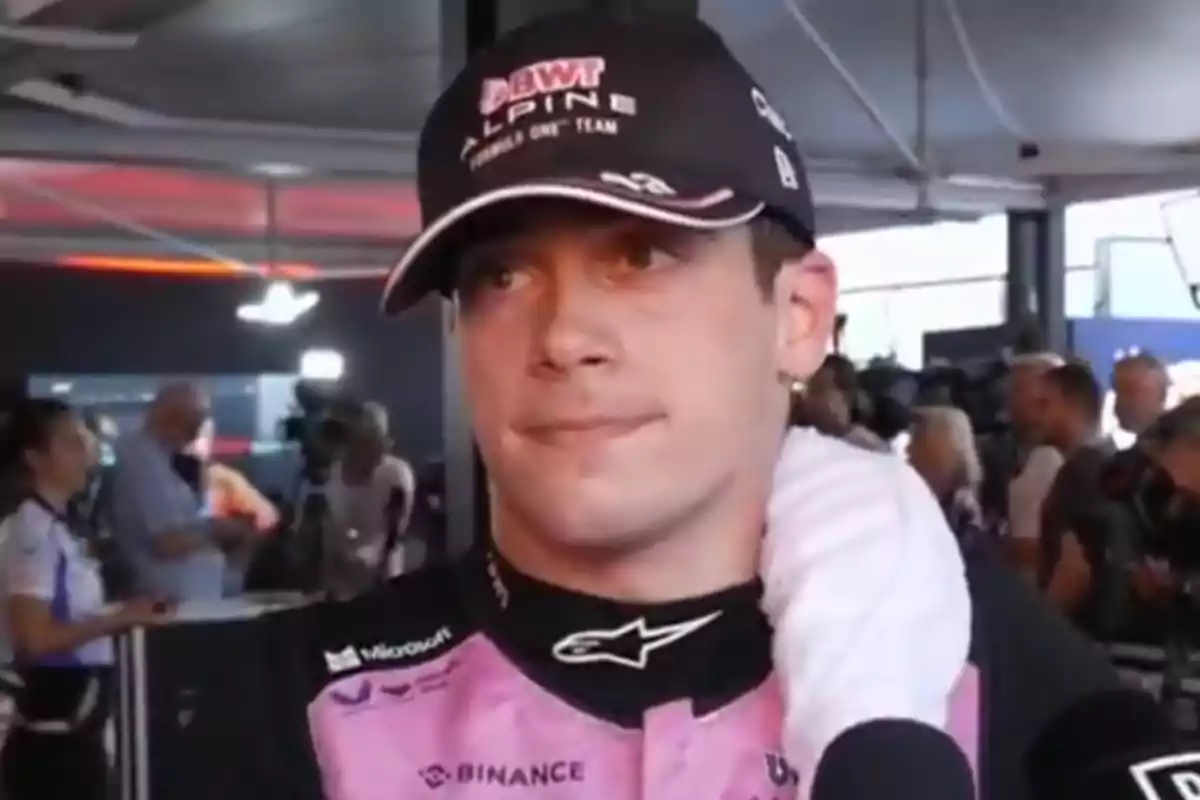 Piloto de automovilismo con uniforme rosa y gorra negra durante una entrevista en el paddock rodeado de periodistas y cámaras