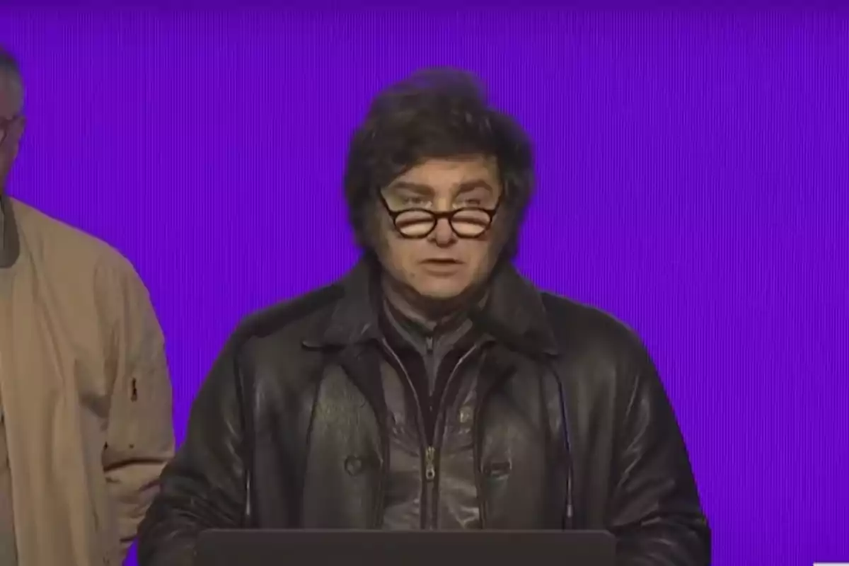Man with glasses and a black leather jacket speaking in front of a purple background