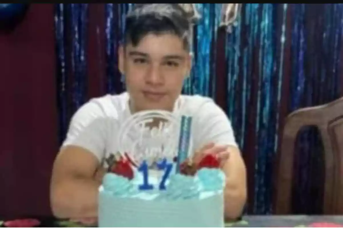 Joven celebrando su cumpleaños número 17 con un pastel azul decorado con fresas y velas, sentado frente a una cortina de tiras brillantes. Joven celebrando su cumpleaños número 17 con un pastel azul decorado con fresas y velas, sentado frente a una cortina de tiras brillantes.