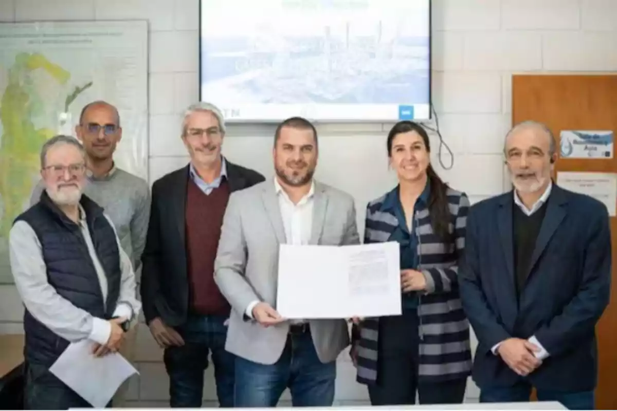 Seis personas posan sonrientes en una oficina mientras una de ellas sostiene un documento frente a una pantalla y un mapa en la pared. Seis personas posan sonrientes en una oficina mientras una de ellas sostiene un documento frente a una pantalla y un mapa en la pared.