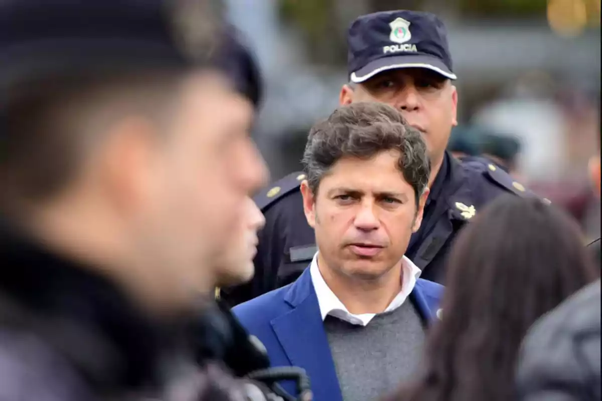Un hombre de cabello corto y saco azul está rodeado de policías con uniforme oscuro en un espacio al aire libre.
