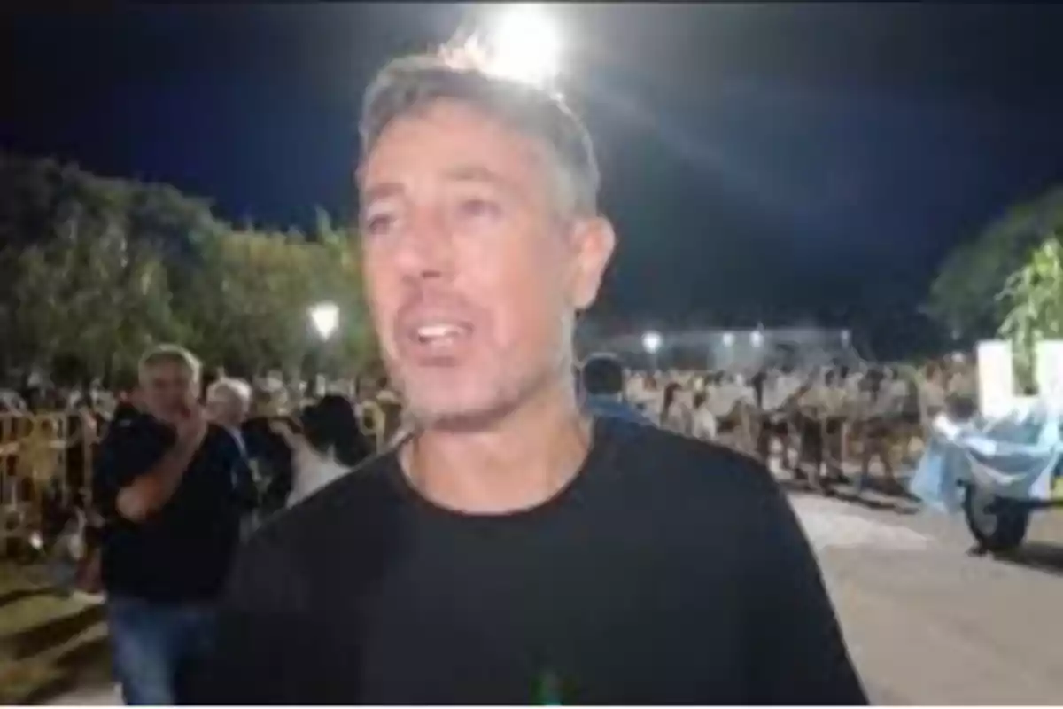Hombre de cabello corto y canoso con camiseta oscura en un evento al aire libre durante la noche con muchas personas al fondo Hombre de cabello corto y canoso con camiseta oscura en un evento al aire libre durante la noche con muchas personas al fondo