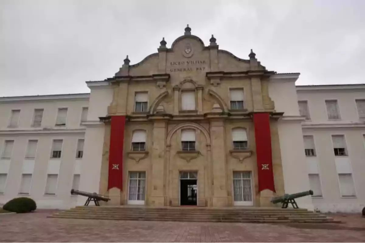 Fachada principal de un edificio histórico con columnas, dos cañones antiguos en la entrada y dos banderas rojas verticales a los costados