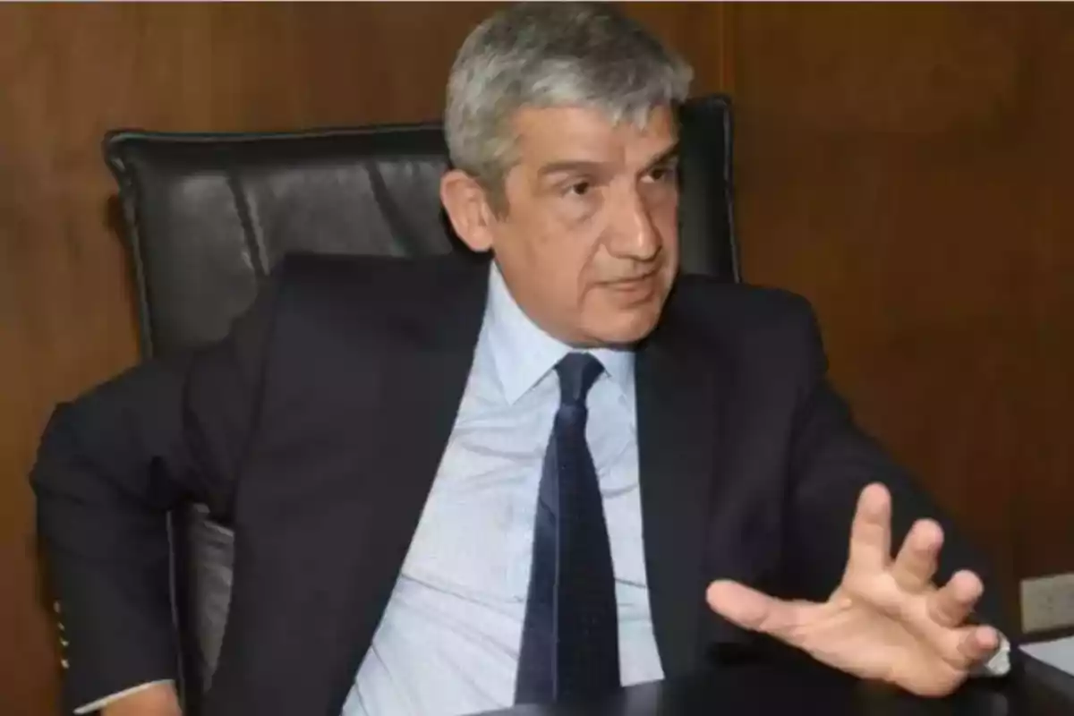 Hombre de cabello canoso con traje oscuro y corbata sentado en una silla de oficina haciendo un gesto con la mano mientras conversa Hombre de cabello canoso con traje oscuro y corbata sentado en una silla de oficina haciendo un gesto con la mano mientras conversa