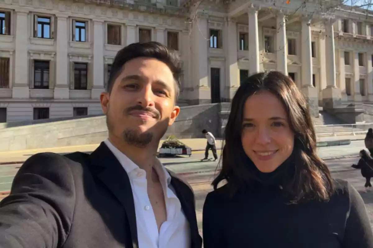 Two people smile while taking a selfie in front of an old building with white columns on a sunny day.