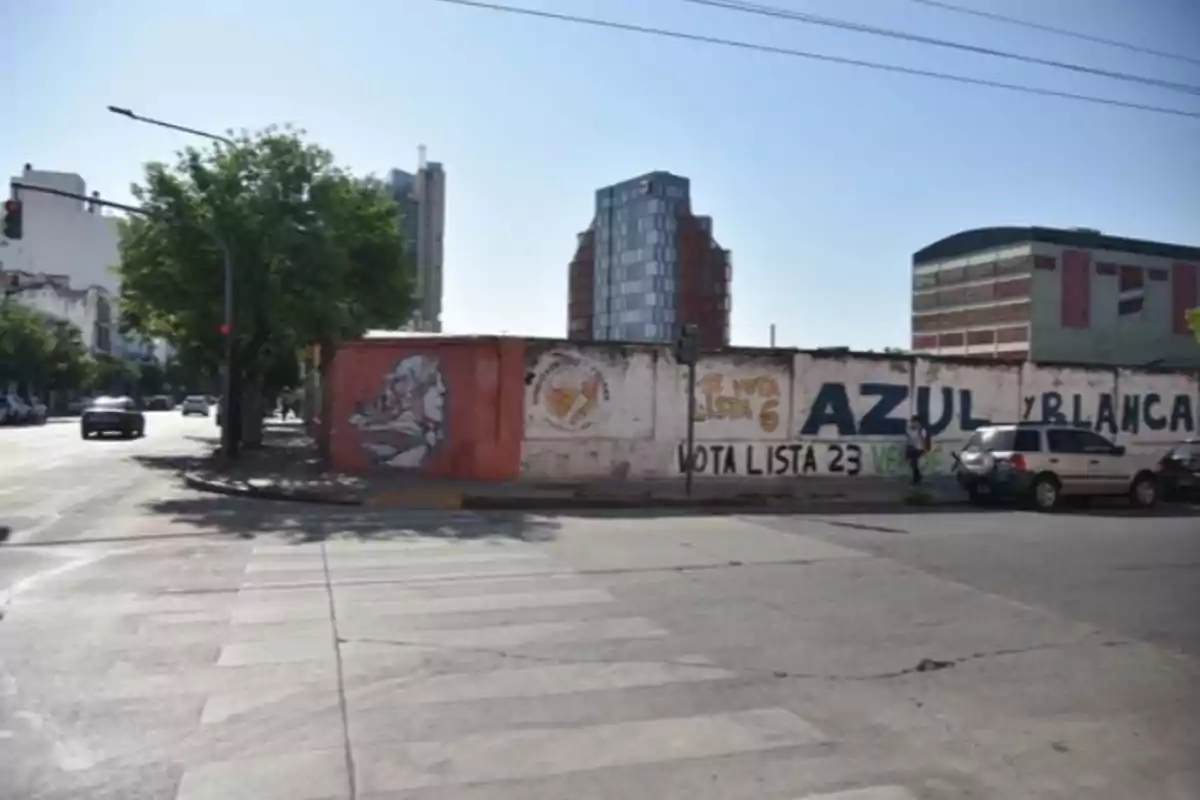 Street intersection in a city with a mural of a woman and political propaganda painted on a wall, some parked cars, and buildings in the background