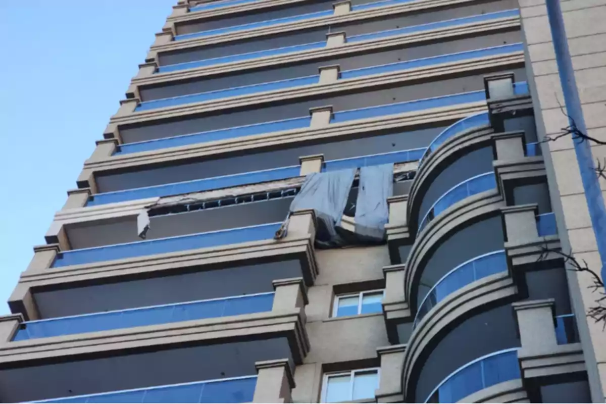 Tall building with blue glass balconies and a gray tarp hanging from one of the upper floors where structural damage can be seen.