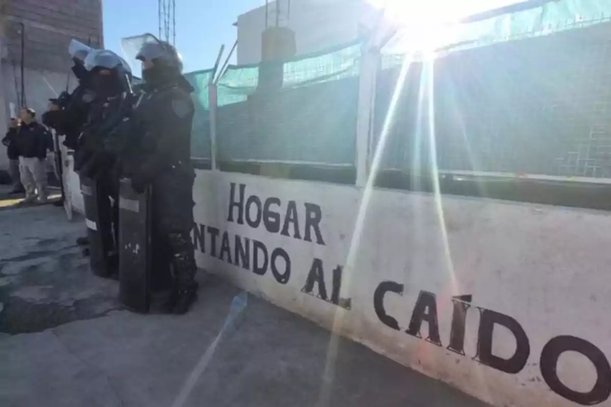 Several police officers in riot gear are standing next to a white wall with a painted message, and the sun is shining brightly on the scene.