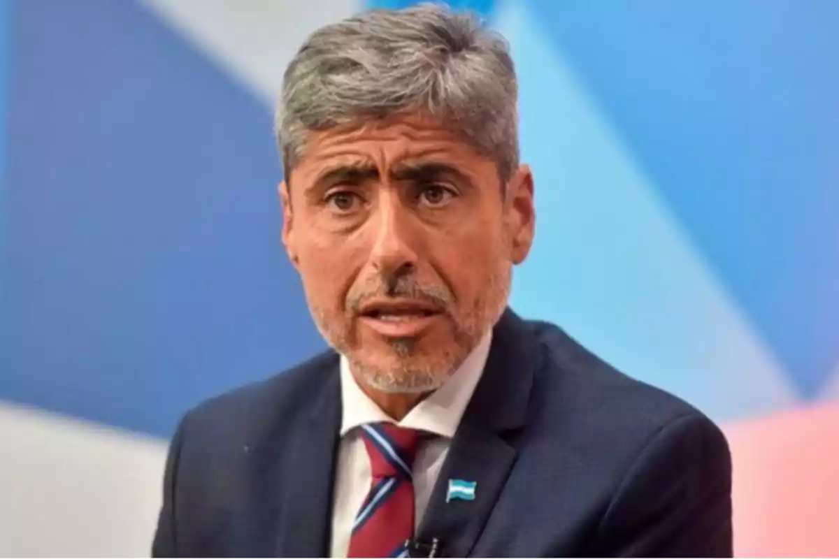 Gray-haired man with a short beard wearing a dark suit with a tie and an Argentine rosette against a blue background. Gray-haired man with a short beard wearing a dark suit with a tie and an Argentine rosette against a blue background.