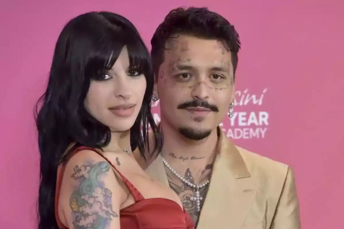 Una pareja posa frente a un fondo rosa, ambos tienen tatuajes visibles y visten ropa elegante.