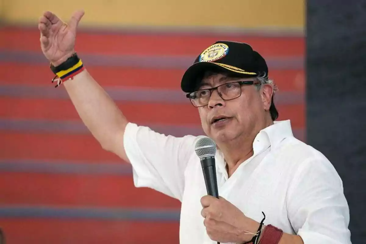 Man with glasses and a black cap speaking into a microphone and raising one hand in front of a red background Man with glasses and a black cap speaking into a microphone and raising one hand in front of a red background