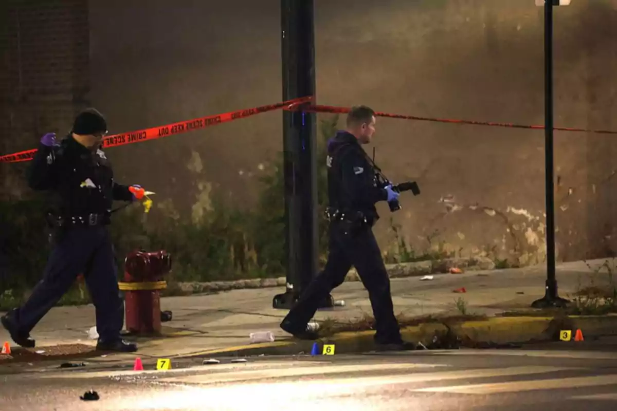Dos policías investigan una escena acordonada en una calle durante la noche, con conos de evidencia en el suelo y cinta amarilla de precaución Dos policías investigan una escena acordonada en una calle durante la noche, con conos de evidencia en el suelo y cinta amarilla de precaución