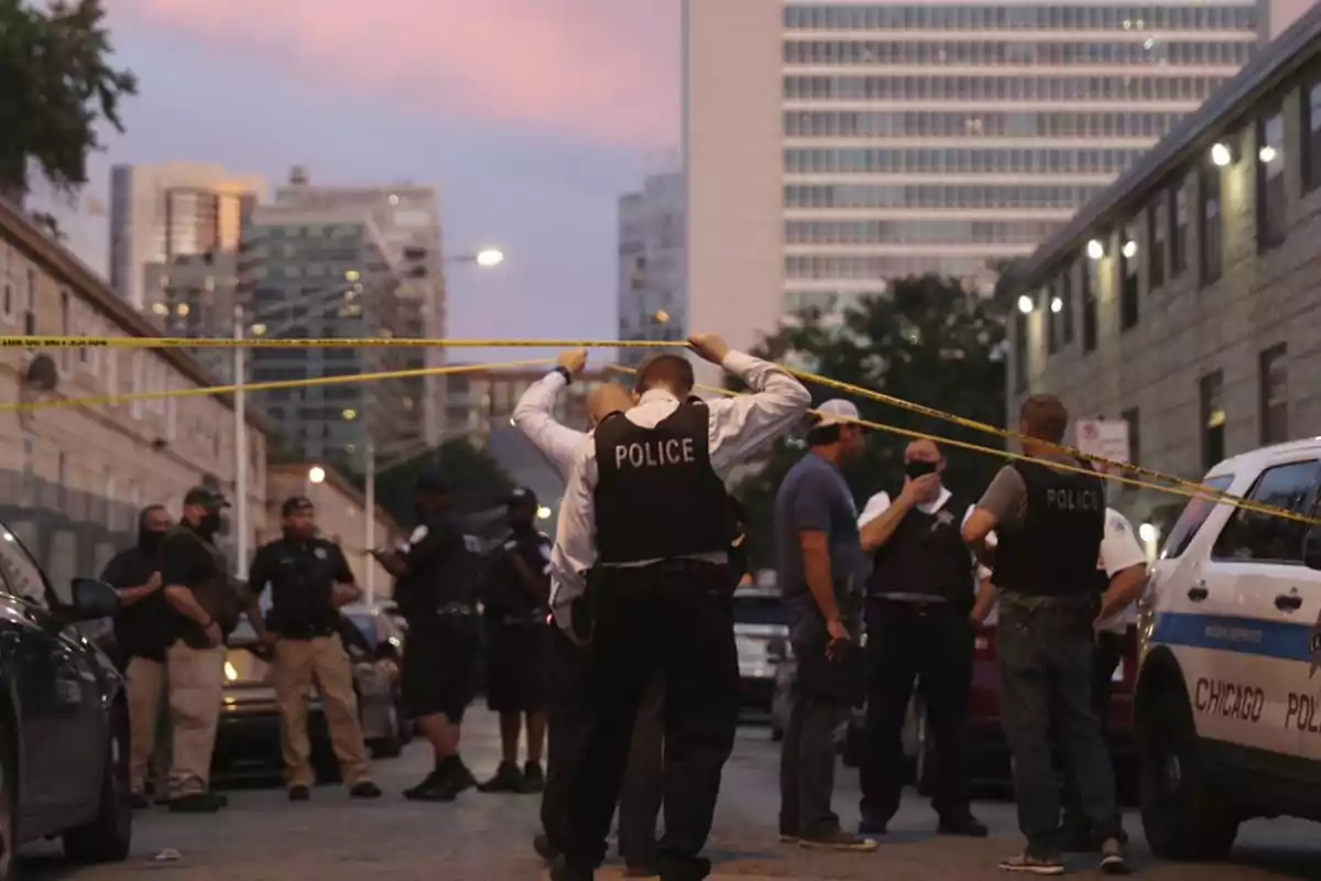 Several police officers and people are on a street cordoned off with yellow tape in an urban area at sunset.