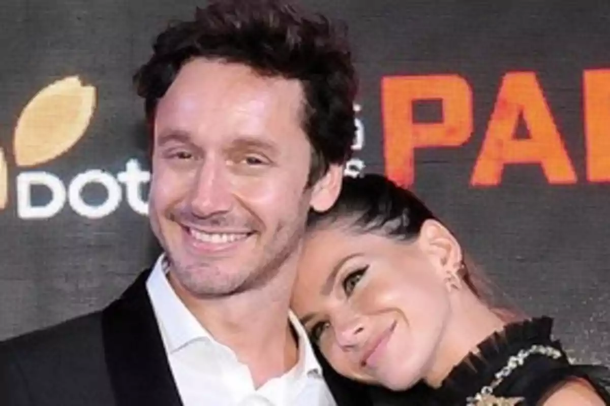 A couple smiles while posing together at a fancy event with a background of orange and white letters. A couple smiles while posing together at a fancy event with a background of orange and white letters.
