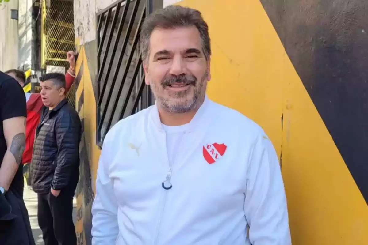 Short-haired, bearded man wearing a white Independiente jacket smiling in front of a yellow and black wall Short-haired, bearded man wearing a white Independiente jacket smiling in front of a yellow and black wall