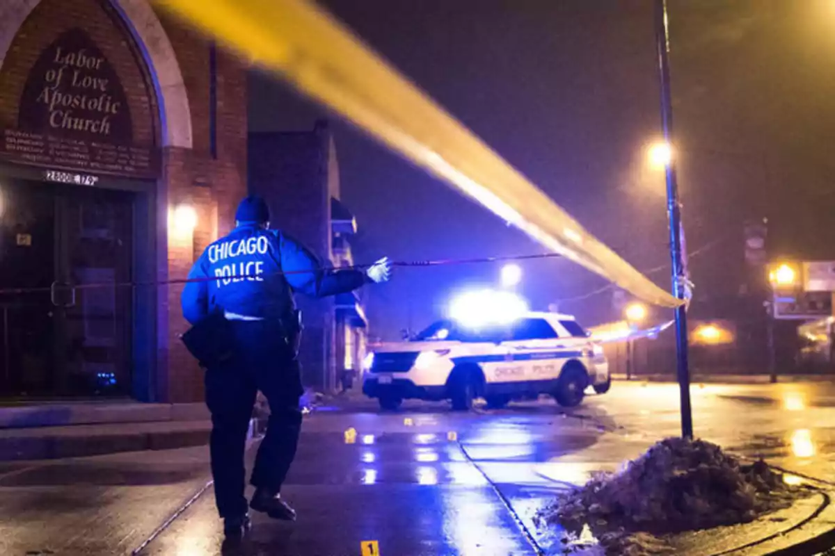 Un policía de Chicago coloca cinta de seguridad en una escena de crimen durante la noche frente a una iglesia, con un patrullero y luces encendidas al fondo