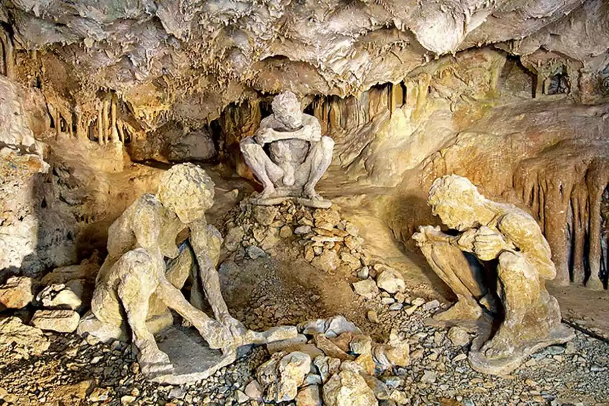Three prehistoric-looking human figures made of stone are inside a cave surrounded by stalactites and rocks. Three prehistoric-looking human figures made of stone are inside a cave surrounded by stalactites and rocks.