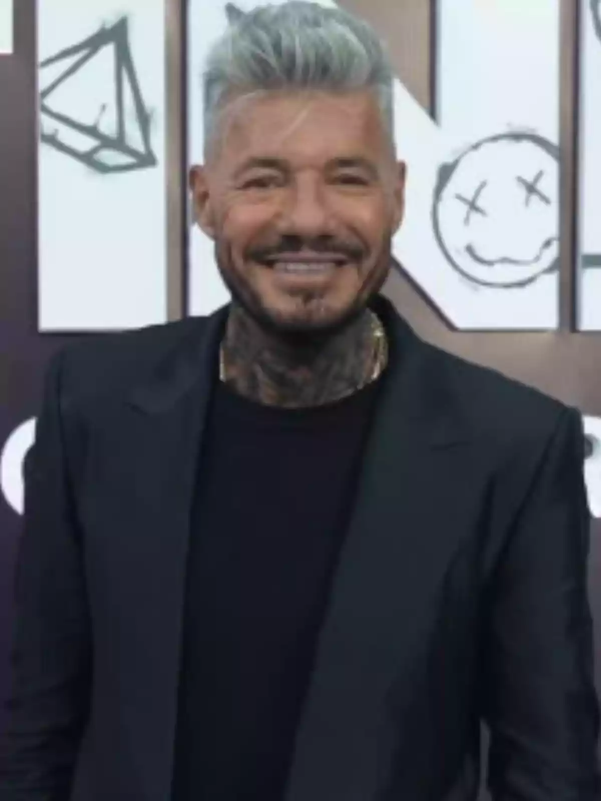 Gray-haired, bearded man wearing a black jacket and dark t-jersey, smiling in front of a background with black and white drawings Gray-haired, bearded man wearing a black jacket and dark t-jersey, smiling in front of a background with black and white drawings