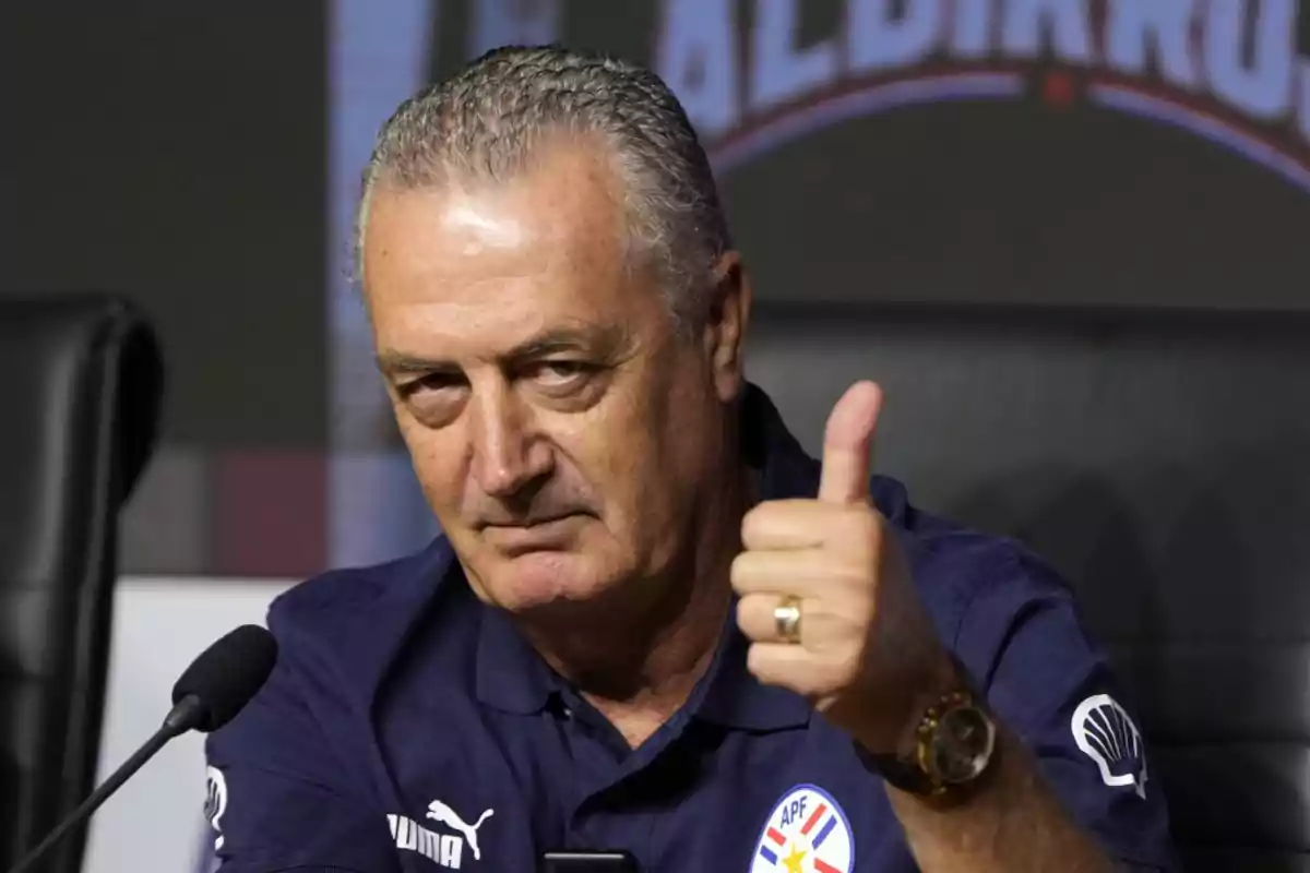 Gray-haired man wearing a blue Paraguay national team jersey giving a thumbs up in approval during a press conference.