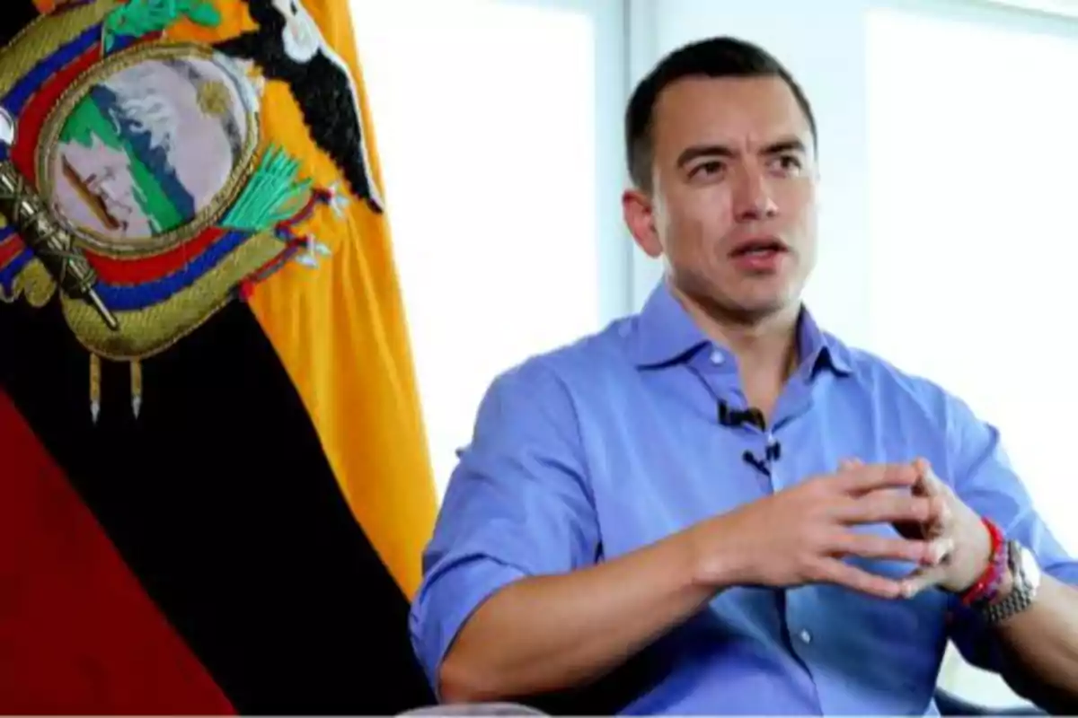 Man in a blue jersey speaking and gesturing with his hands next to an Ecuadorian flag in a well-lit indoor setting