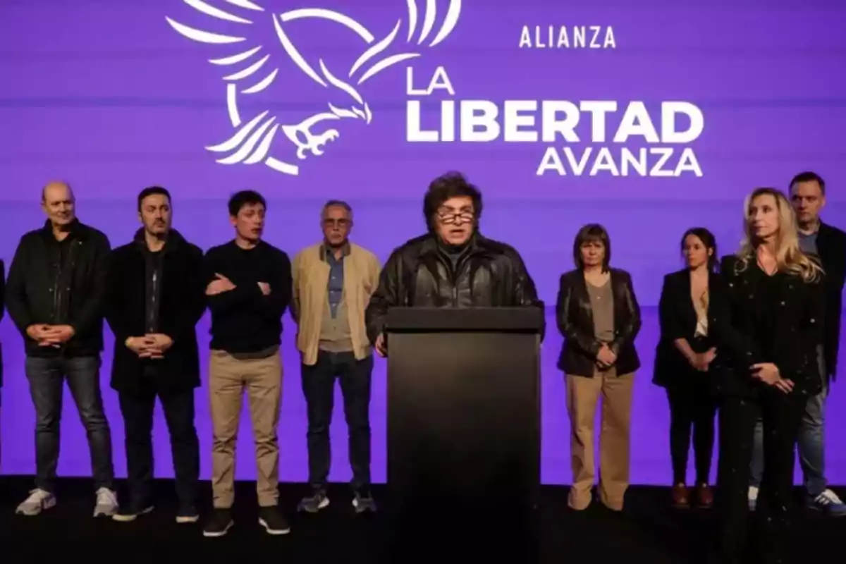 A group of people is standing on a stage with a purple background, and a man in the center is speaking from a lectern; behind him, the logo of an eagle and the text "La Libertad Avanza" can be seen.