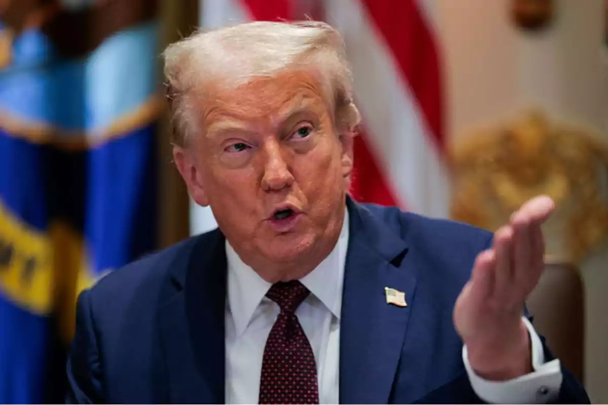 Hombre de cabello rubio con traje azul y corbata roja gesticulando con la mano mientras habla en un entorno oficial con banderas al fondo Hombre de cabello rubio con traje azul y corbata roja gesticulando con la mano mientras habla en un entorno oficial con banderas al fondo