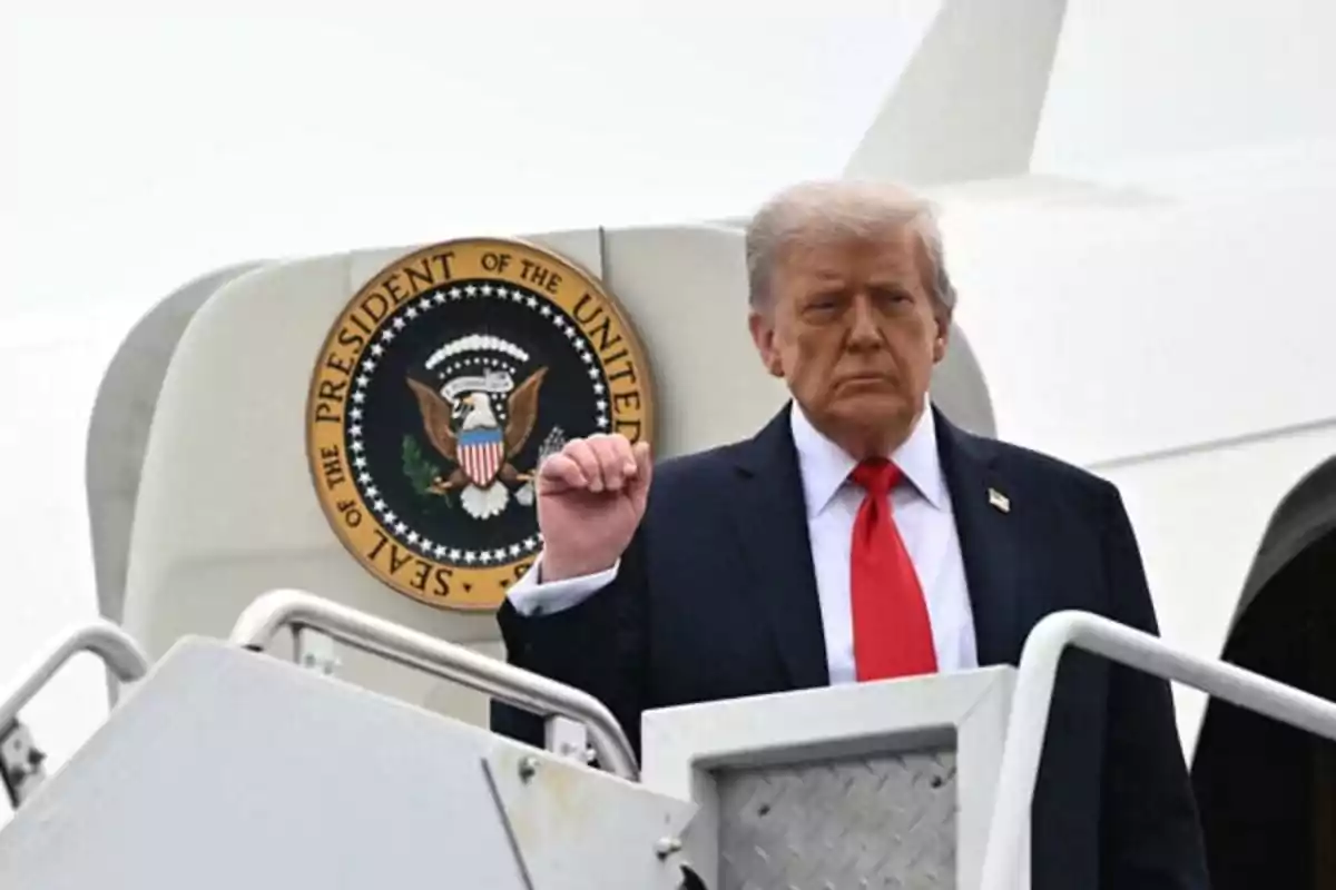 Blond-haired man in a dark suit and red tie climbing the stairs of a presidential airplane with the United States seal in the background