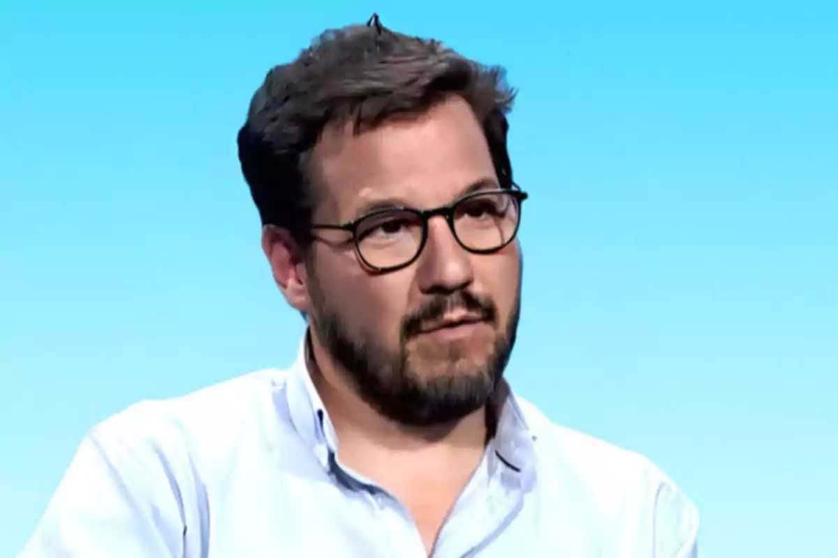 Man with a beard and glasses wearing a light jersey against a blue background