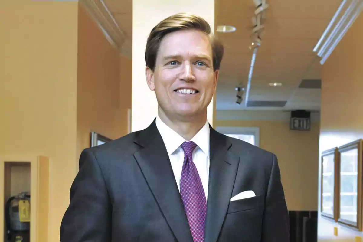 Hombre de cabello rubio con traje oscuro y corbata morada sonriendo en un pasillo iluminado Hombre de cabello rubio con traje oscuro y corbata morada sonriendo en un pasillo iluminado
