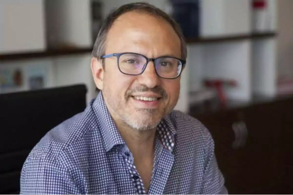 Hombre de mediana edad con gafas y camisa a cuadros sentado en una oficina sonriendo Hombre de mediana edad con gafas y camisa a cuadros sentado en una oficina sonriendo